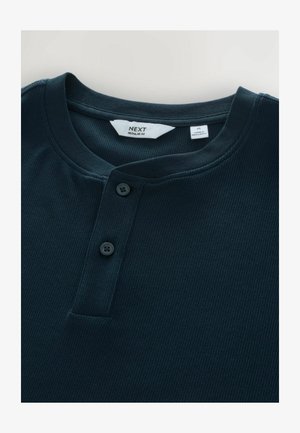Tummansinisessä ribattussa henley-paidassa on pyöreä pääntie, kaksi mustaa nappia ja etiketti, joka osoittaa koon keskikoko, NEXT-merkkinen.