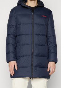 Chaleco acolchado de color azul marino con capucha, con diseño acolchado, cierre de cremallera y logo rojo "HUGO" en el pecho. Material texturizado e insulated.