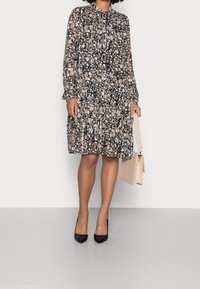 Blommönstrad klänning med långa ärmar, en nyckelhålsringning och en flerdelad kjol, kombinerad med en beige handväska och svarta högklackade skor.