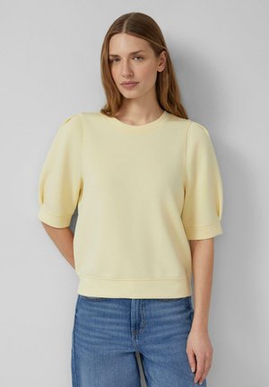 Femme aux longs cheveux brun clair portant un haut jaune pâle à manches courtes bouffantes et un jean bleu, debout devant un fond gris clair uni.