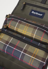 Interieur van de Barbour rugzak met olijfgroen stof en twee ritssluitingen in geplaidpatroon in groene, marineblauwe, rode, witte en gele tinten.