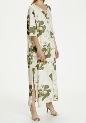 Kvinde iført en lang, løs, cremefarvet kjole med store grønne blomsterprint og sideslidser, kombineret med beige, sandal-stropper med høje hæle.