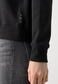 Sudadera negra con puños y dobladillo acanalados. Presenta costuras decorativas en verde, plata y negro en la costura lateral. La tela parece suave.