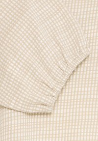 Helle beigefarbene Stoff mit kariertem Muster, versehen mit Raffdetails an den Ärmeln und einer weichen, strukturierten Oberfläche.