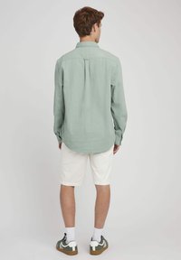 Chemise à manches longues vert clair en tissu texturé, avec des poignets à boutons et un pli au dos. Assortie à un short blanc et des baskets multicolores.