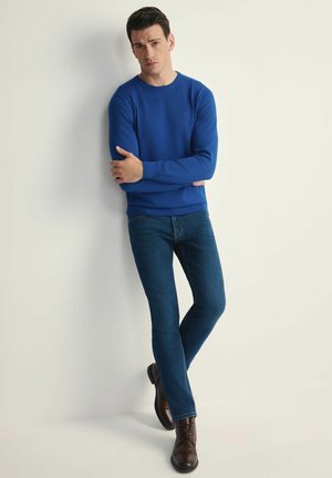 Niebieski sweter w okrągłym dekolcie, połączony z ciemnoniebieskimi dżinsami i brązowymi butami na sznurówki; stoi na jasnym tle.