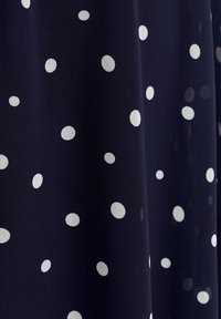 Tissu bleu marine foncé avec de petits pois blancs espacés de manière irrégulière, montrant des plis doux et une texture.