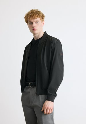 Fiatal férfi göndör vörös hajjal, fekete bomber dzsekit, fekete inget, szürke nadrágot és fekete övet visel, egyszerű fehér háttér előtt.