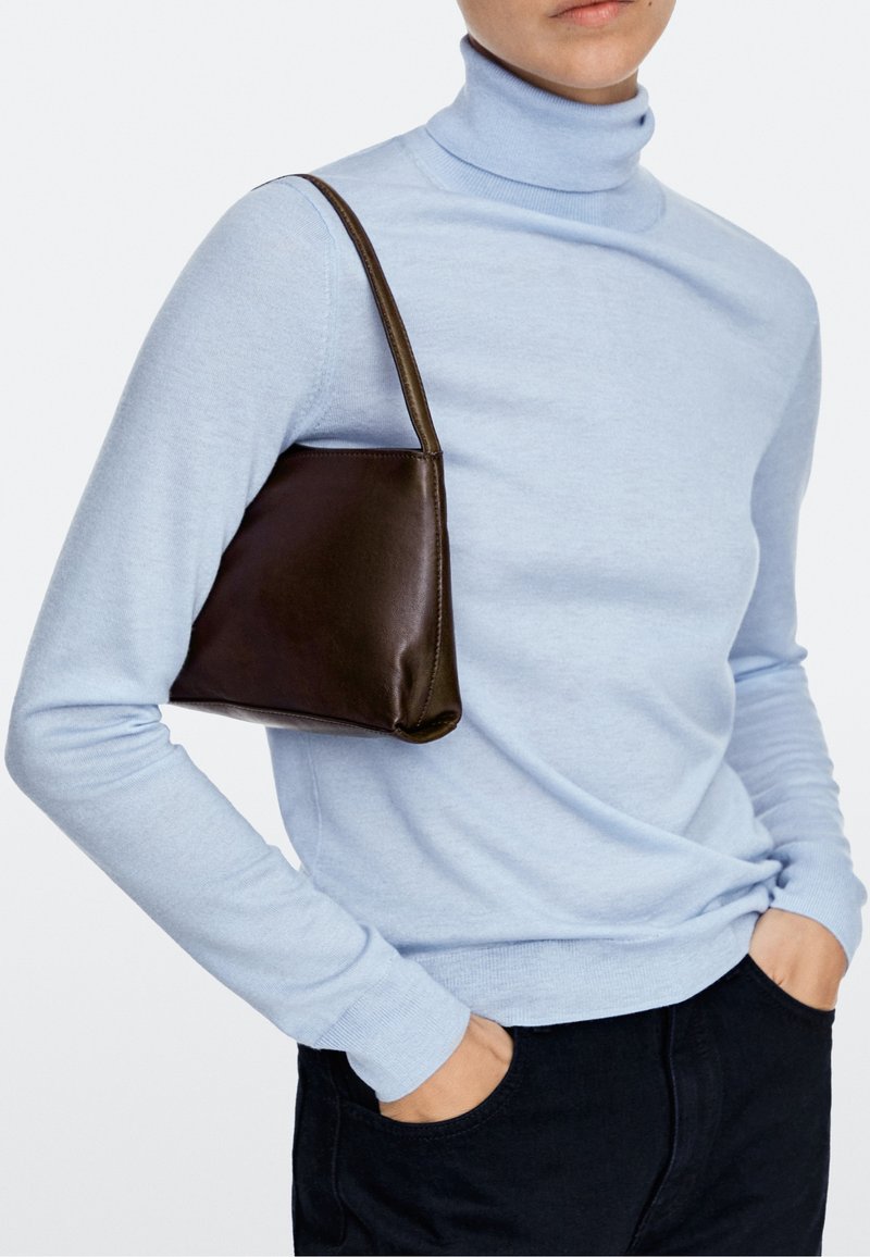 Person wearing a light blue turtleneck sweater and black pants holds a small dark brown leather shoulder bag on their left shoulder.