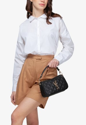 Donna che indossa una camicia bianca con bottoni e pantaloncini marroni tiene in mano una borsa nera trapuntata con logo e dettagli dorati, impugnandola dal manico superiore.