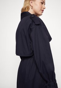 Trench coat blu navy con colletto strutturato, dettagli con bottoni sulle spalle e legatura regolabile in vita. Tessuto morbido con vestibilità su misura.