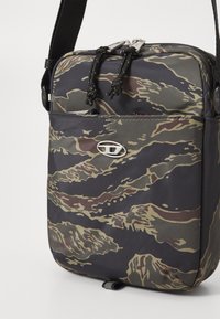 Borsa a tracolla compatta con motivo camouflage verde scuro e marrone, tasca frontale con zip, tracolla nera regolabile ed emblema logo argentato.