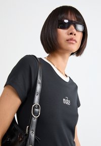 Fekete póló fehér gallérral, hímzett "nike" logóval. Fekete válltáskával és sötét napszemüveggel viselve.