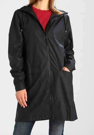 Femme portant un imperméable noir à capuche avec fermeture éclair avant et grandes poches, par-dessus un haut rouge et un jean bleu.