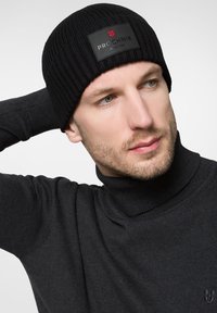 Czarna dziana czapka z logotypem. Dopasowany krój z prążkowaną teksturą. Nosi się z ciemnym swetrem typu golf, odsłaniając fragment skóry.