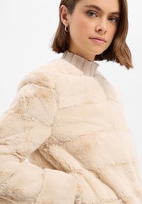 Beiger Pullover aus Kunstfell mit strukturierten horizontalen Streifen und einem beigen, gerippten Kragen. Der Stoff wirkt weich und flauschig, mit einer lockeren Passform.