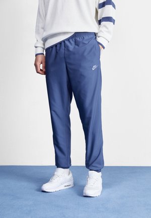 Persona che indossa pantaloni da tuta Nike blu, scarpe da ginnastica bianche e un maglione bianco con strisce blu, in piedi su un pavimento con moquette blu.