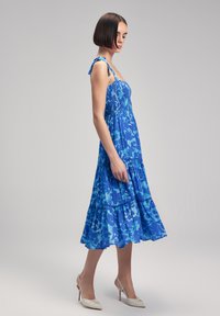Abito midi a fiori blu con corpetto arricc-biè, spalline regolabili e orlo arricciato. Tacchi bianchi lucidi completano l'outfit.