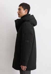 Schwarze isolierte Parka mit Kapuze, ausgestattet mit einem Reißverschluss, Seitentaschen und einem Patch am linken Ärmel. Glatte Stofftextur.
