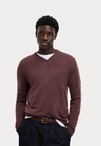 Suéter burdeos de cuello en V en un suave tejido de punto, presenta mangas largas y puños acanalados. Llevado sobre una camisa blanca, combinado con pantalones oscuros.