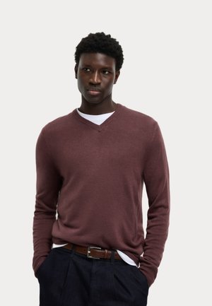 Pullover a V in bordeaux, realizzato in un morbido knit, con maniche lunghe e polsini a coste. Indossato sopra una camicia bianca, abbinato a pantaloni scuri.