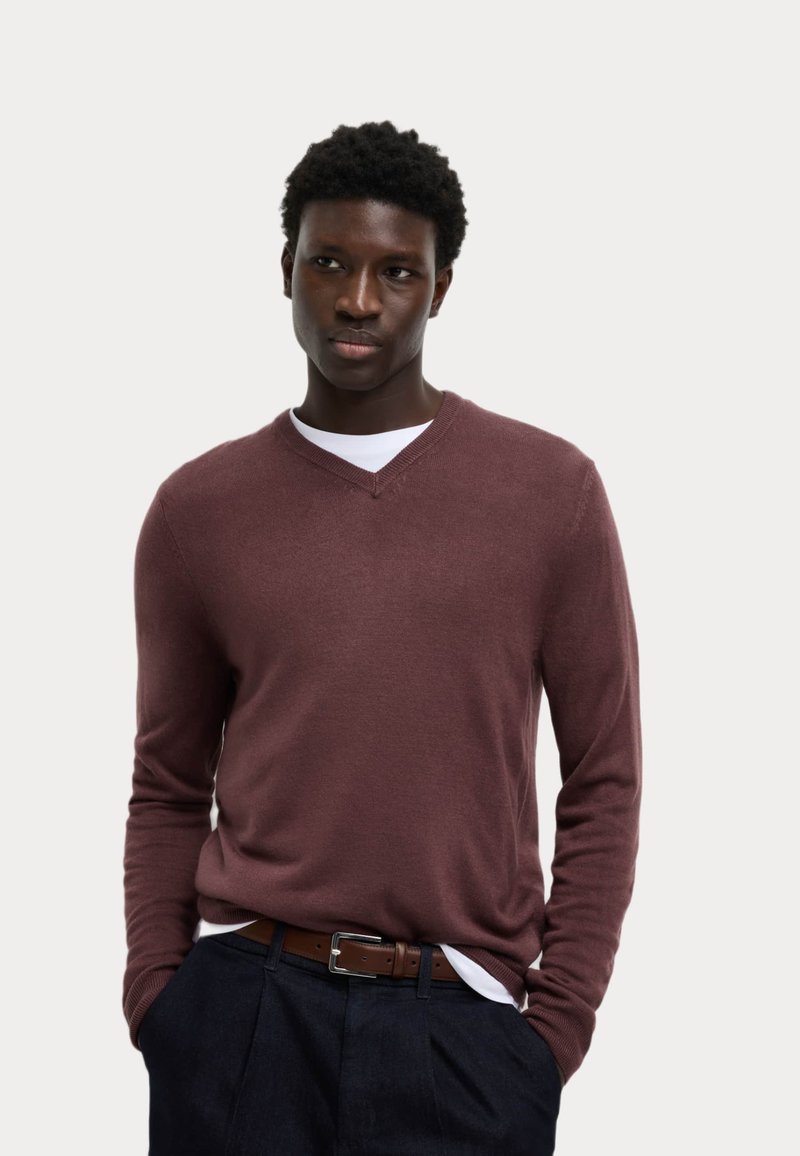 Suéter burdeos de cuello en V en un suave tejido de punto, presenta mangas largas y puños acanalados. Llevado sobre una camisa blanca, combinado con pantalones oscuros.