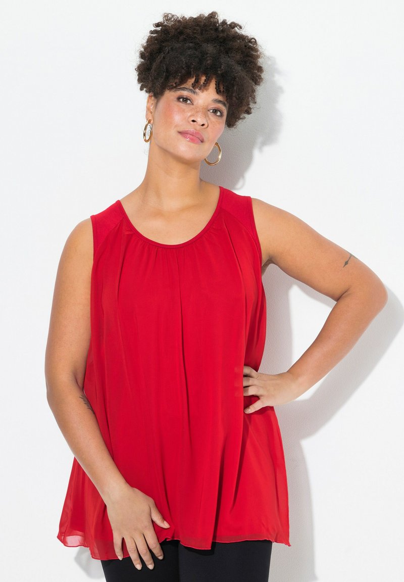 Top rosso senza maniche realizzato in un tessuto leggero e fluido, con scollatura rotonda e morbidi plissè, abbinato a pantaloni neri. Orecchini a cerchio dorati visibili.