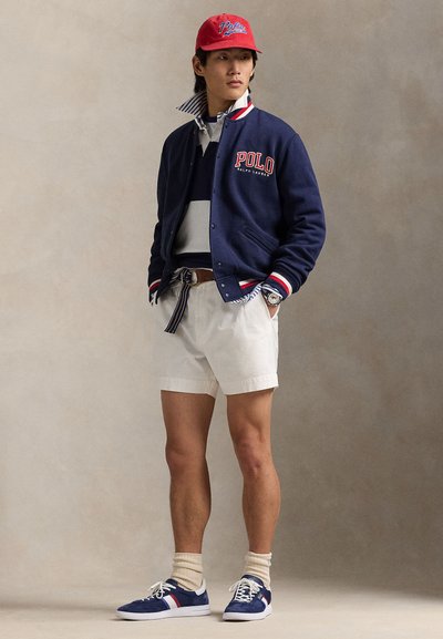 Mladi moški, ki nosi rdečo kapo, mornarsko modro šolsko jakno z logotipom "Polo", črtasto srajco, bele kratke hlače, črtast pas in modre superge, stoji pred enobarvnim ozadjem.