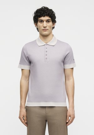 Νέος άνδρας με σγουρά σκούρα μαλλιά που φοράει ανοιχτό μωβ κοντομάνικο πόλο με μπεζ γιακά και μανσέτες, στέκεται μπροστά σε απλό φόντο.