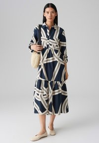 Navy and cream patterned shirt dress with long sleeves, button-down collar, tiered skirt, and a woven bag. Smooth fabric.