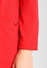 Manche de blazer rouge avec un revers roulé, en tissu lisse. Comprend un bouton argenté en accent et une couture détaillée le long de la couture.