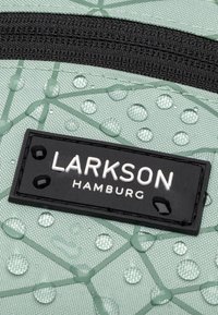 Zaino verde chiaro in materiale con superficie texturizzata, con zip nera e patch del logo. Gocce d'acqua visibili sul tessuto.