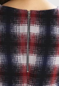 Tissu à carreaux noir, rouge et blanc avec une finition texturée. Présente une fermeture éclair en métal argenté à l'arrière, située au centre.