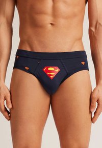 Caleçons pour hommes bleu marine en tissu lisse, ornés d'un grand logo Superman rouge et jaune à l'avant, avec des logos plus petits dispersés autour.