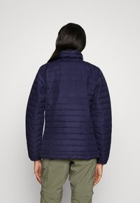 Granatowa kurtka puchowa z wysokim kołnierzem, poziomym pikowanym wzorem i gładką teksturą. Noszona na oliwkowymi spodniami, odsłaniająca część rękawa.