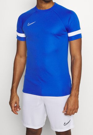 Mężczyzna w niebieskiej sportowej koszulce z krótkim rękawem, białym paskiem na rękawach i białych szortach, oba z logo Nike.