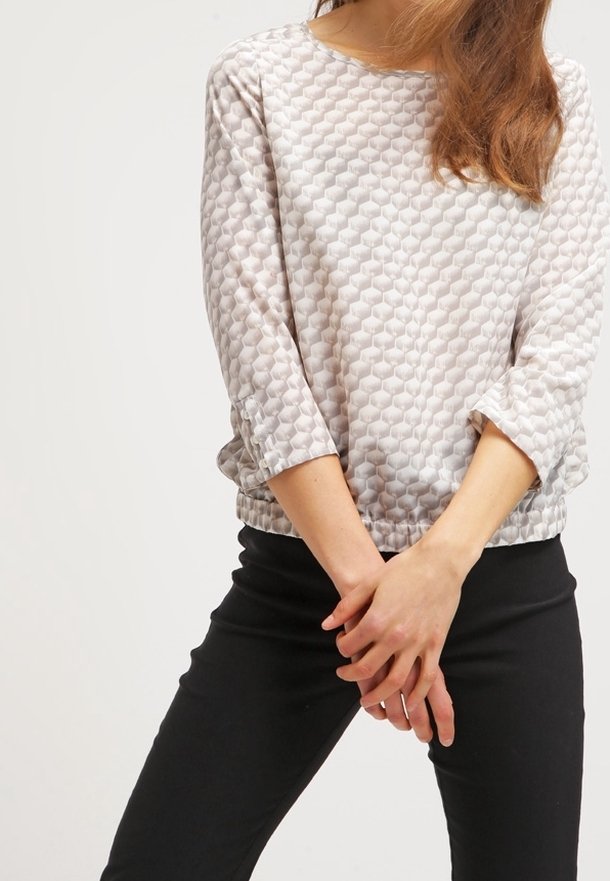Blouse légère à motifs dans un doux gris avec un design hexagonal texturé, dotée de manches 3/4 et d'une coupe décontractée. Portée avec un pantalon noir.