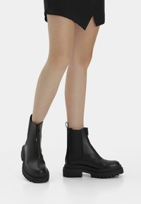 Černé kožené kotníkové boty s robustní podešví, vybavené elastickými bočními panely a předním zipem. Texturovaný vzorek pro lepší přilnavost a odolnost.