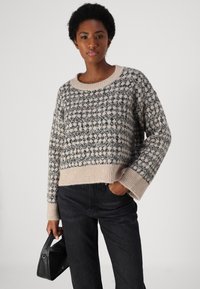 Pull en tricot beige avec un motif agrémenté d'accents bleu foncé. Coupe courte, manches larges et poignets côtelés, associé à un jean foncé.
