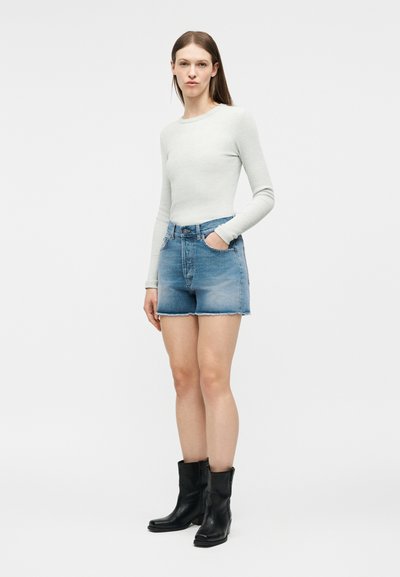 Γυναίκα με μακριά καστανά μαλλιά φοράει ανοιχτό γκρι μακρυμάνικη μπλούζα, μπλε τζιν σορτς με ρεβέρ και μαύρες δερμάτινες μπότες μέχρι τη μέση της γάμπας, όρθια.