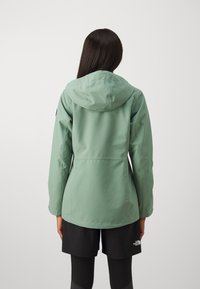 Veste imperméable vert clair avec capuche, présentant une coupe droite, des poignets élastiques et un tissu lisse. Logo noir sur la manche.
