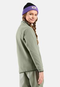 Veste en polaire verte claire avec un col haut, associée à un pantalon vert clair ; le mannequin porte un bonnet en tricot noir et violet avec un logo.
