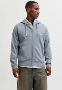 Sweat à capuche gris zippé avec une poche frontale, des cordons de serrage et des poignets côtelés. Porté par-dessus une chemise à carreaux avec col.
