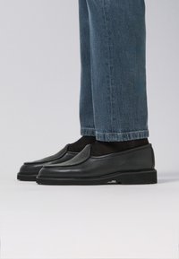 Mocassini in pelle nera con finitura texturizzata, design slip-on e suola piatta in gomma, abbinati a jeans di denim blu.