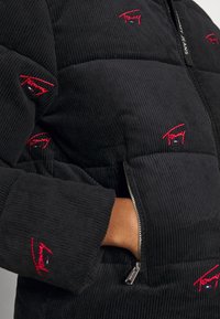 Czarna kurtka puchowa z sztruksu, z czerwonymi wyszywanymi logo Tommy Jeans, kieszeniami na zamek i ściągaczami na rękawach. Wyraźna tekstura i detale wzornicze.