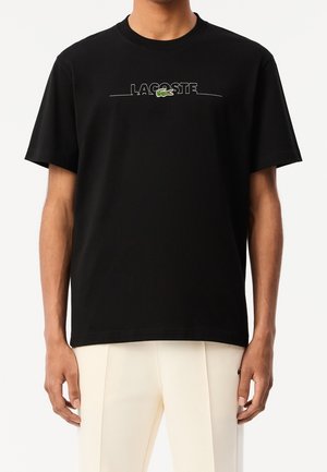 Homme portant un t-shirt noir Lacoste avec un logo brodé blanc et un emblème de crocodile vert, associé à un pantalon blanc cassé.