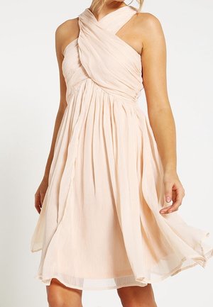 Robe rose pâle sans manches, longueur genou, avec un col halter croisé et une jupe fluide froncée, portée par une personne debout.