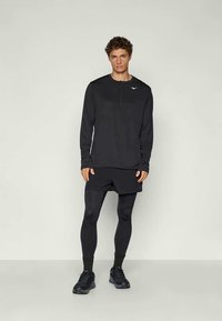 Svart långärmad träningströja med dragkedja framtill, i kombination med svarta shorts och leggings, iförd svarta löparskor.