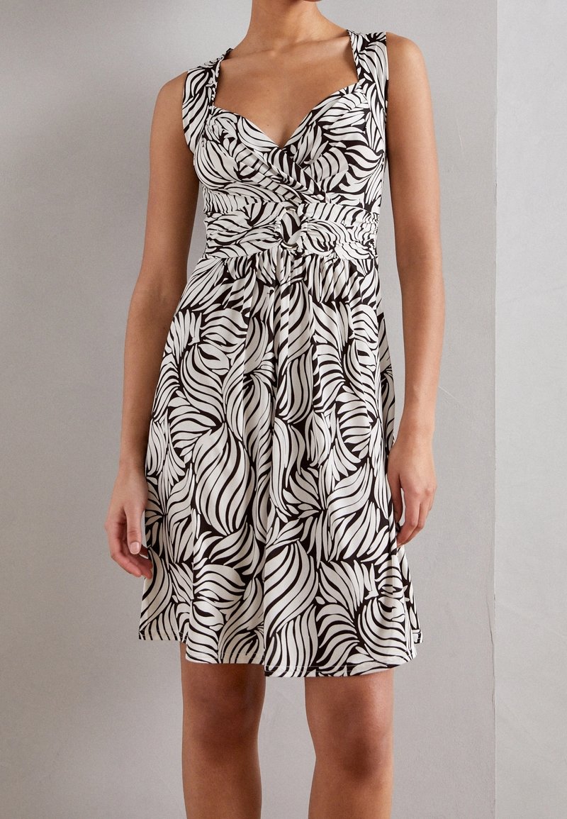Femme portant une robe sans manches à motifs noirs et blancs avec une taille froncée et un décolleté en V, debout contre un mur uni gris clair.