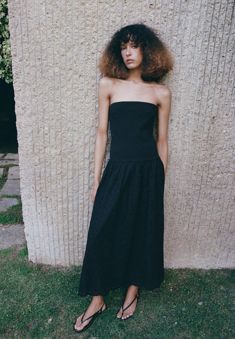 Jeune femme aux cheveux bouclés portant une robe noire sans bretelles et des sandales noires, debout contre un mur de pierre texturé sur l'herbe.
